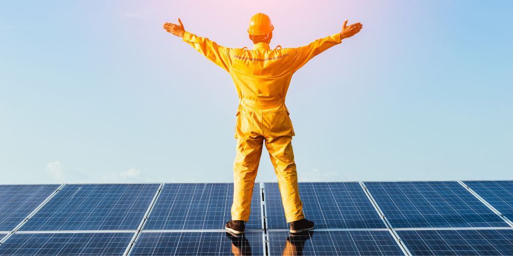 A person in an orange jumpsuit stands on solar panel,s with their arms spread wide